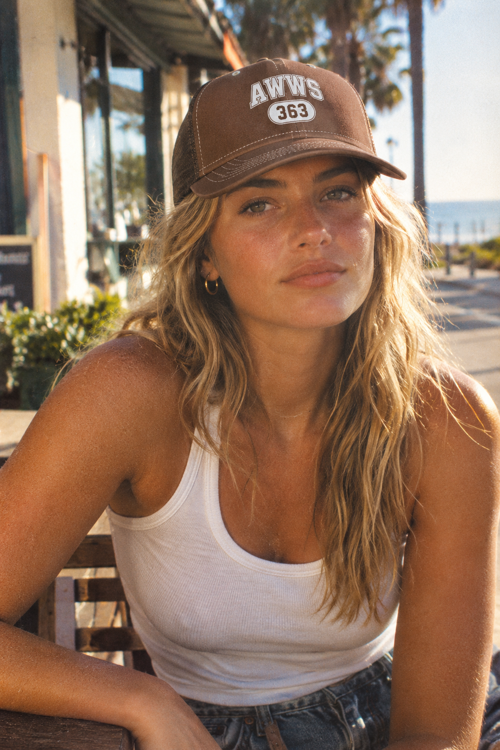 A woman wearing a brown trucker-style cap with 'AWWS 365' printed on the front, sitting outdoors near a storefront with palm trees and the ocean in the background.